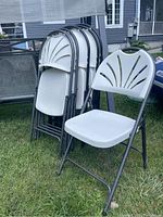 Photo showing six white folding chairs stacked together outdoors on grass, highlighting the molded plastic seat and back design with metal frames.