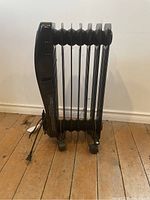 Side view of black electric radiator heater showing heating fins and caster wheels on wooden floor.