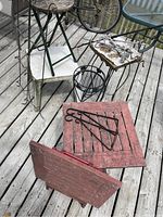 Multiple assorted garden tables and related accessories on a wooden deck including a folding table with marble top, red wooden table, wrought iron and metal stands.