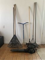 Wide view of five garden tools including three metal rakes, one long-handled cultivator and an electric leaf blower, placed upright and lying on a wooden floor against a white wall.
