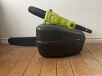 Green small electric hedge trimmer placed on a closed black plastic carrying case for the chainsaw.