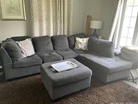 View of gray L-shaped sectional couch and matching ottoman in living room with brown rug and window curtains.