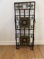 Front view of the metal folding magazine rack featuring decorative tiles and scrollwork