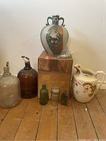 Overview of assorted vases and bottles included in the lot, showing large decorative ceramic vase, large white ceramic pitcher, two large glass bottles, and three small glass containers on wooden floor.