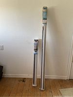 Two zebra blinds in original clear plastic packaging leaning against wall on hardwood floor, showing length difference