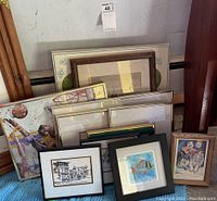 Lot of 11 assorted framed artworks of varying sizes stacked on floor against wall.
