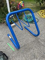 Front-angle view of blue metal bike rack with triangular frame, showing scratches and outdoor setting with garden hose and flower decorations nearby.