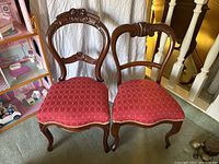 Pair of vintage wooden chairs with red diamond-patterned upholstery on seats, one with carved top rail and floral details, the other with simpler curved backrest, positioned side by side on carpet.