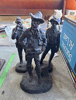 Photo shows three dark cowboy statues standing on floor with visible details of clothing and poses.