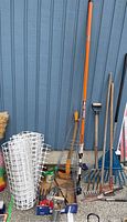 Full view of various gardening tools including axe, rakes, tomato stakes, and netting against a blue wall on concrete surface.