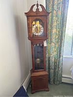 Full view of the grandmother clock showing overall shape, size, and placement near a window with floral curtains.