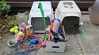 Photo showing two plastic Petmate Kennel Cab pet kennels with metal doors, various pet supplies including tennis balls, leash, harness, ball tosser, stake, cracked pet food bowl, cords and miscellaneous items in white box on concrete patio.