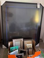 Large black rectangular picture frame with white protective corner covers placed behind a box of smaller assorted picture frames of various colors and styles.