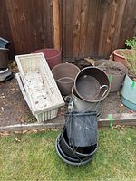 Photo showing a mix of round plastic pots, metal bucket-style pots with handles, and two long rectangular planter boxes stacked outside by a wooden fence.