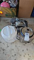 View of white plastic rice cooker, Rival food slicer, and stainless steel Cuisinart rice cooker/steamer on concrete floor in garage.