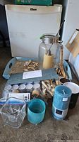 Overview of the lot showing a light blue sand tray with sand, driftwood pieces, various candles including a large pillar candle in a glass lantern, blue and black container candles, tealight candles, and glass candle holders in front of a white cooler.