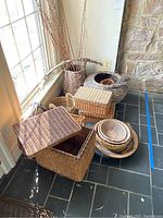 Various woven baskets, boxes, wreaths, bowls, and vase arranged on floor near window and stone wall.