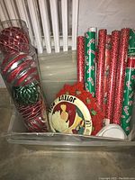 Photo showing multiple rolls of Christmas wrapping paper, vintage metal plaques, large Christmas ball ornaments in plastic container.