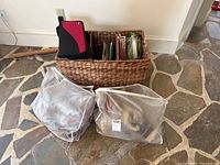 Full view of the wicker basket with knitting needles and two large plastic bags of yarn in front on stone floor.