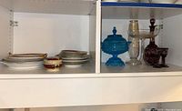 Shelf view showing the blue glass covered dish, giant martini glass, clear glass pitcher, decanter with stopper and some plates and bowls to the side.