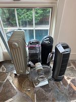 View showing two black tower heaters, one white oil-filled radiator heater, and a small oscillating desk fan in front, all on a tiled floor near a glass door.
