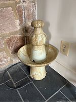 Full view of ceramic fountain on tiled floor near stone wall and electric outlet. Shows entire fountain and surrounding area.
