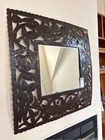 Front view of the square Haitian metal framed mirror hanging on a wall showing floral and bird cut-out metal design around the glass.