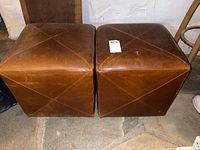 Pair of brown leather cube hassocks placed side by side on stone floor against white wall, showing wear and scratches on surface.