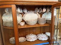 Display of the entire set inside a wooden china cabinet, showing cups, saucers, plates, and covered casseroles.