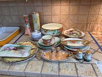 Full view of all Limoges, Noritake, Bavarian china serving plates, teacup, tray, salt and pepper shakers, and vases arranged on countertop.
