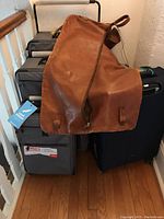 World's Lightest luggage set of three soft gray rolling suitcases with branded tags and handles, and a brown leather suit carrier draped over luggage handles.