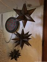 Photo of three perforated metal hanging stars in various sizes hanging against a wall with a dart board visible in the background.