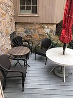 Outdoor patio setup showing six dark brown plastic wicker chairs around two tables, one larger white metal table and one smaller brown wicker table, with a closed red checked patio umbrella anchored in a metal stand.