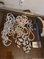 Six pearl necklaces in different shades and one pair of pearl stud earrings in jewelry box displayed on a reflective surface.