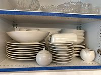 Upper cabinet shelf showing stacks of dinner plates, salad plates, bowls, cups, saucers and sugar bowl