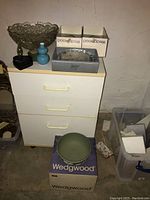 Lot overview: Wedgwood bowl on box, cut glass bowl, blue bud vases, black glass heart trinket box, crystal picture frame and glass candlesticks on white cabinet