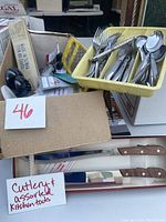 Overall view of the assortment including knives with wooden handles in original packaging, a yellow tray with forks and spoons, and other miscellaneous kitchen tools and cutlery.