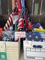 Multiple US flags in various styles, some packaged in plastic, folded, and stacked with foreign flags visible in box along with outdoor thermometers.