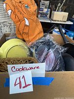 Box with cookware including yellow pot, packaged skillet, orange oven mitt, woven trivets, and black pans