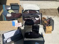 Photo showing multiple pairs of men's dress shoes in boxes (brown and black), shoe rack, and other footwear accessories arranged outside on concrete surface.