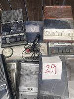Photo showing multiple vintage Panasonic tape recorders, a microphone, and small electronic devices clustered together.
