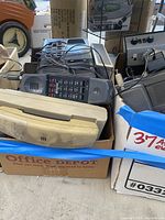 Box containing beige rotary phone handsets, black cordless phone handset with visible keypad, and parts of other phones and electronics
