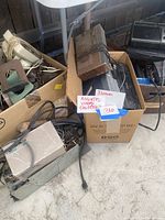 Photo showing several vintage electronic items including a beige rotary telephone, cables, power adapters, and possibly an old turntable or speaker in cardboard boxes.