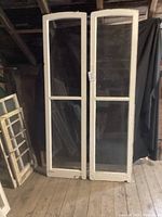 Pair of tall white wooden frame antique windows with clear glass panes, viewed standing upright against a wall