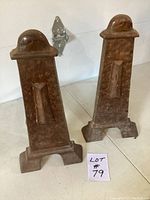 Pair of two heavy, rusted fireplace andirons standing upright on a white surface. Each has a distinct stepped base and domed top.