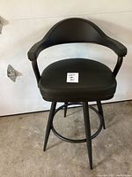 Front view of black leather bar stool showing padded seat and curved backrest with armrests, angled legs and circular footrest ring