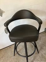 Front view of black leather bar stool showing cushioned seat, curved backrest with armrests, and black four-legged base.