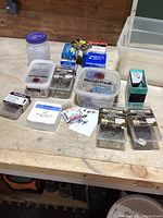 Overview of lot showing multiple plastic containers and boxes of nails and staples of various sizes and brands on a wooden surface.