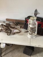Photo shows multiple rusty metal pulleys, a wooden beam, a vintage metal lantern, and a red and black tool bag on a white table.