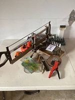 Full view of the miscellaneous tools on table including bow saw, measuring tapes, protractors, pipe cutter, drawing compass, small hacksaw and other hand tools